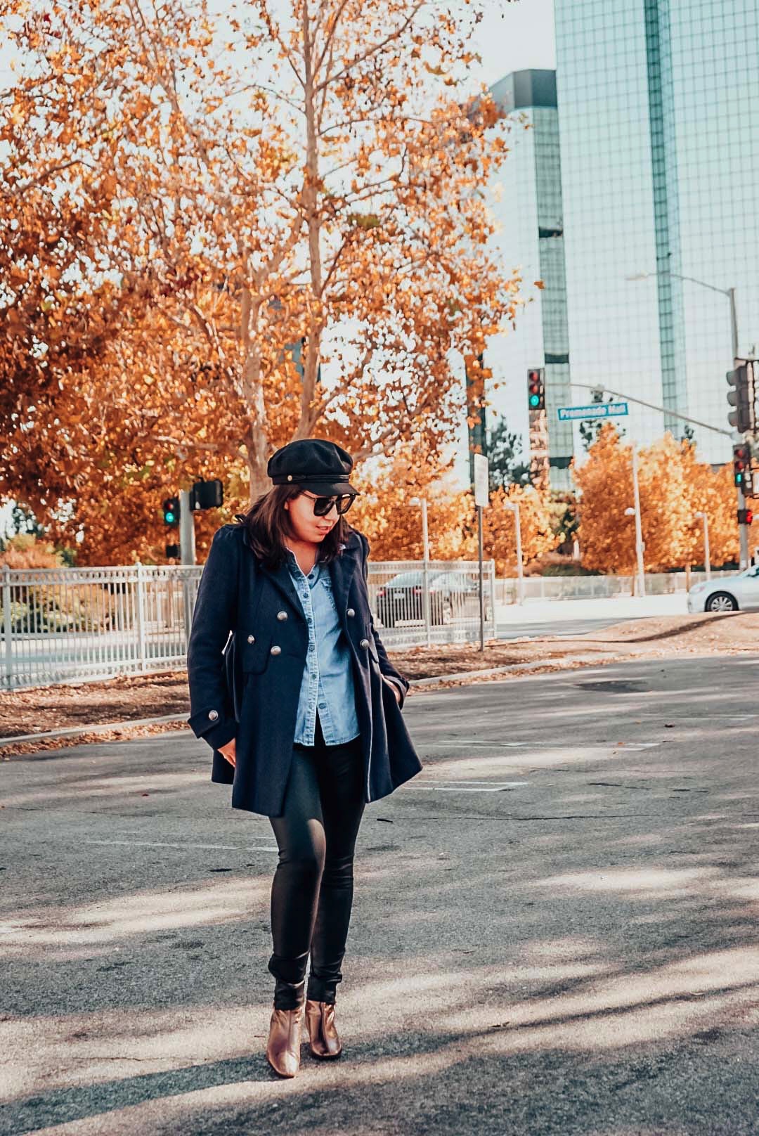 Autumn foliage with a woman wearing navy wool coat, newsboy hat, faux leather leggings, and metallic ankle boots in the city. 