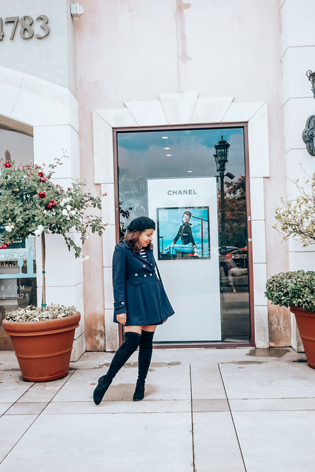 Chanel storefront add with a woman in navy coat, black beret, striped shirt, and over the knee black suede boots.