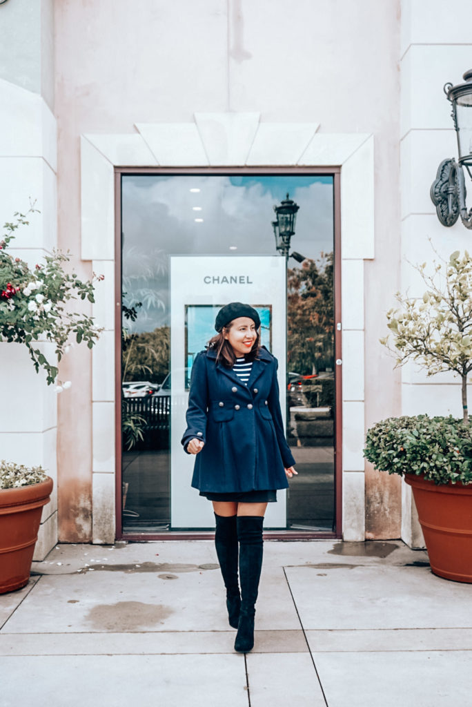 Chanel storefront add with a woman in navy coat, black beret, striped shirt, and over the knee black suede boots.