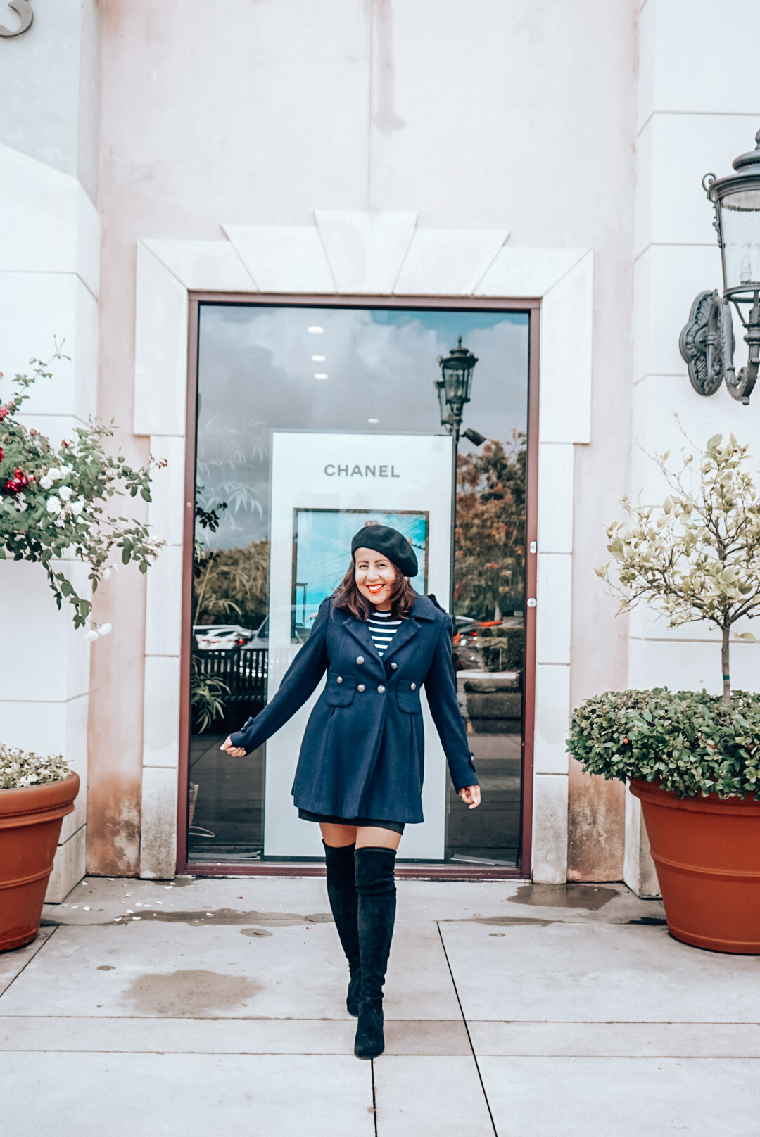 Chanel storefront add with a woman in navy coat, black beret, striped shirt, and over the knee black suede boots.