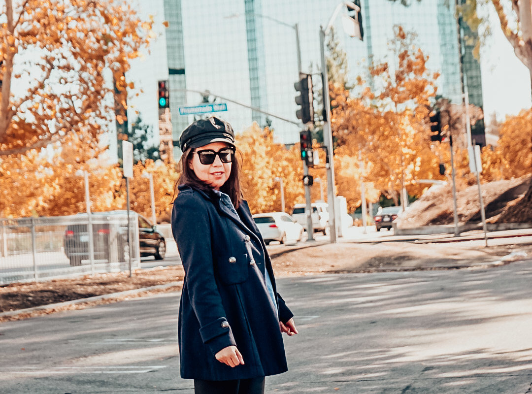 Autumn foliage with a woman wearing navy pea coat, newsboy hat, and metallic ankle boots with faux leather leggings. 