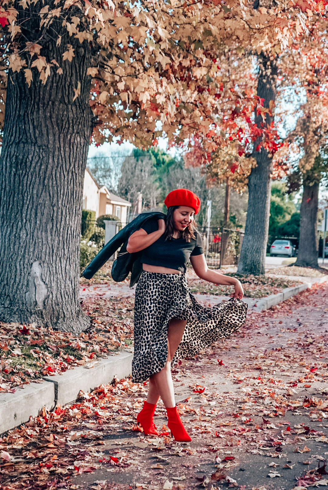 A woman wearing leopard print midi skirt and faux leather jacket with red beret and red ankle boots twirling.