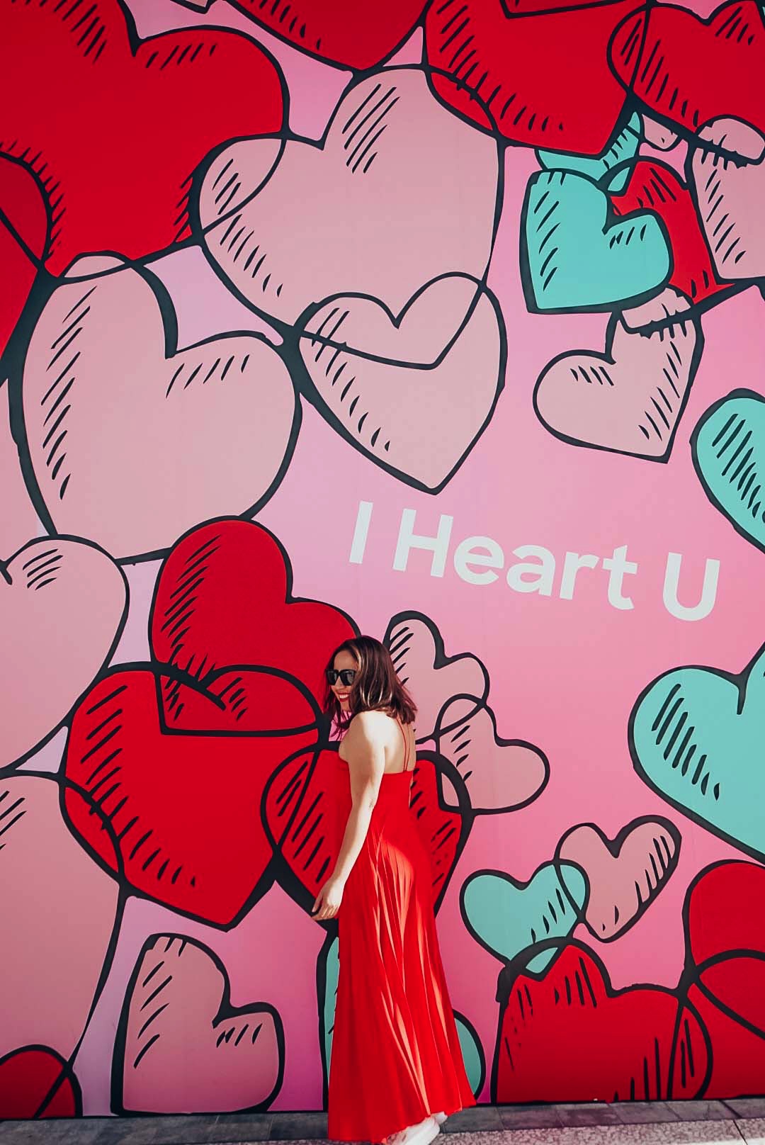 Girl in long red maxi dress strolling in front of a heart mural in Los Angeles. 