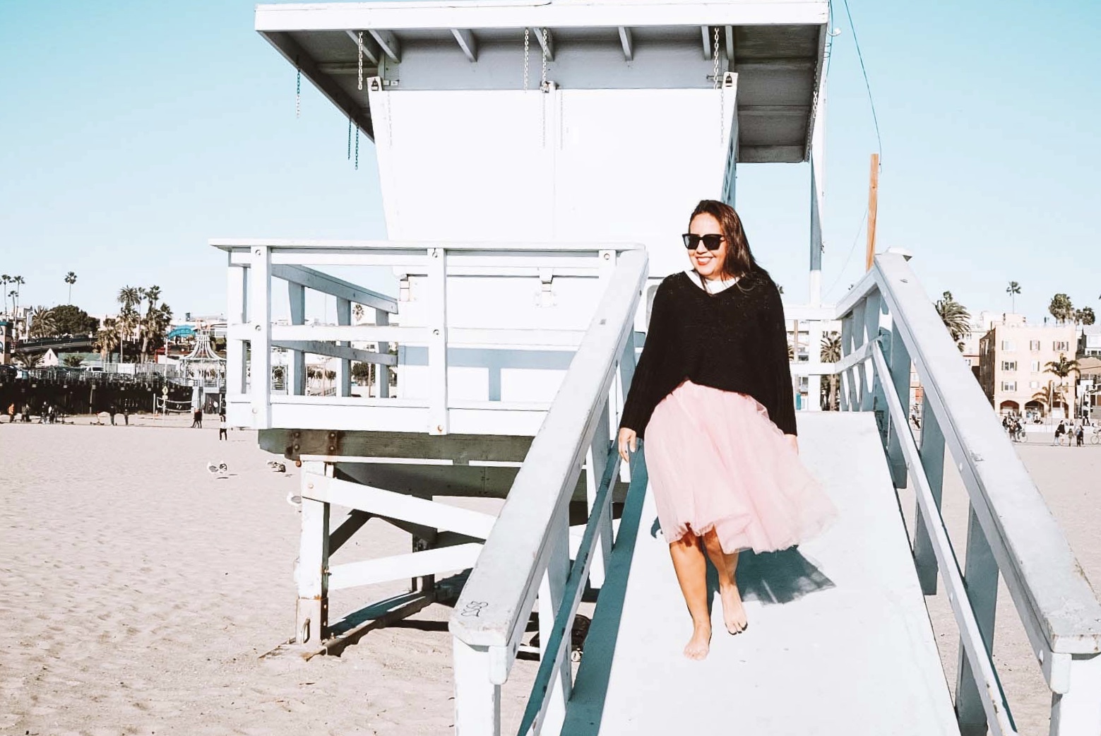 Woman in a pink tutu at the beach smiling wearing sunglasses waling down a lifeguard tower.